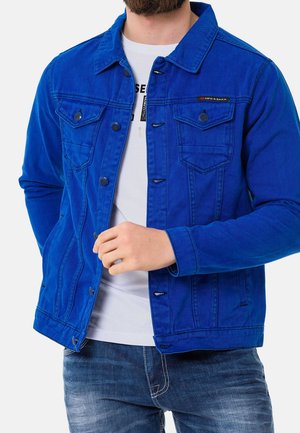Mand iført en lys blå denimjakke over en hvid t-shirt og blå jeans, med den ene hånd, der justerer jakke foran.