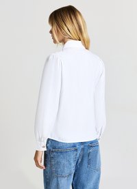 Donna con capelli biondi che indossa una blusa bianca a maniche lunghe e jeans blu, mostrata di spalle su uno sfondo neutro.