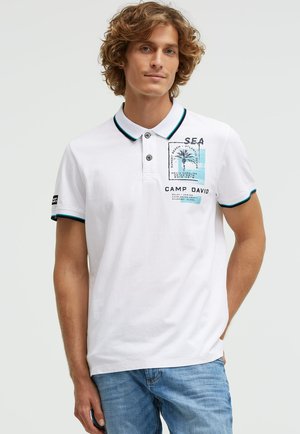 Man wearing white polo shirt with palm tree graphic and text, paired with blue jeans, standing against a plain light background.