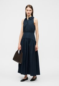 Žena v tmavě modrých nabíraných šatech bez rukávů a s rozšířenou sukní, držící hnědou kabelku a obutá v černých balerínkách, stojící před jednoduchým světlým pozadím.