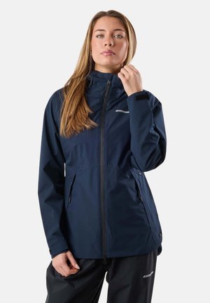 Woman with long hair wearing a navy blue zip-up jacket and black pants, adjusting collar with one hand against a plain background.