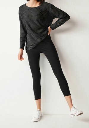 Femme portant un haut à manches longues noir à motifs, des leggings noirs courts et des baskets blanches, posant contre un mur blanc.