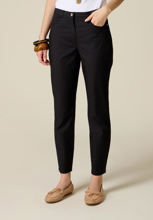 Kvinde iført sorte slim-fit bukser, beige mokkasiner, hvid top og chunky armbånd på venstre håndled, stående mod en ensfarvet baggrund.