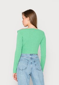 Sweater curto canelado em verde brilhante com mangas longas e design fora do ombro, combinado com umas calças de ganga de cintura alta em azul claro.