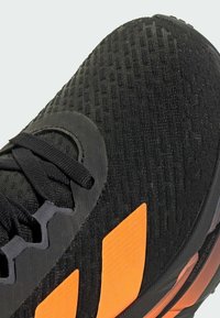 Black athletic shoe with textured mesh upper, featuring orange accents and laces. Includes a patterned finish and a rubber sole with branding.