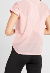 Camiseta deportiva de color rosa claro con mangas cortas, un panel trasero transparente y un corte relajado, combinada con leggings negros.