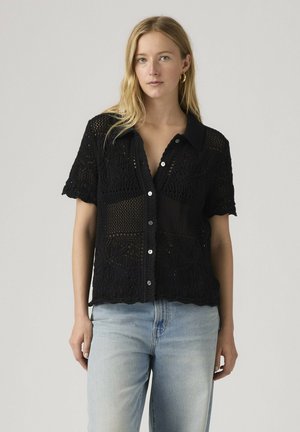 Vrouw draagt een zwart gehaakt kortermet overhemd met knopen en lichtblauwe spijkerbroek, staand tegen een effen lichtgrijze achtergrond.