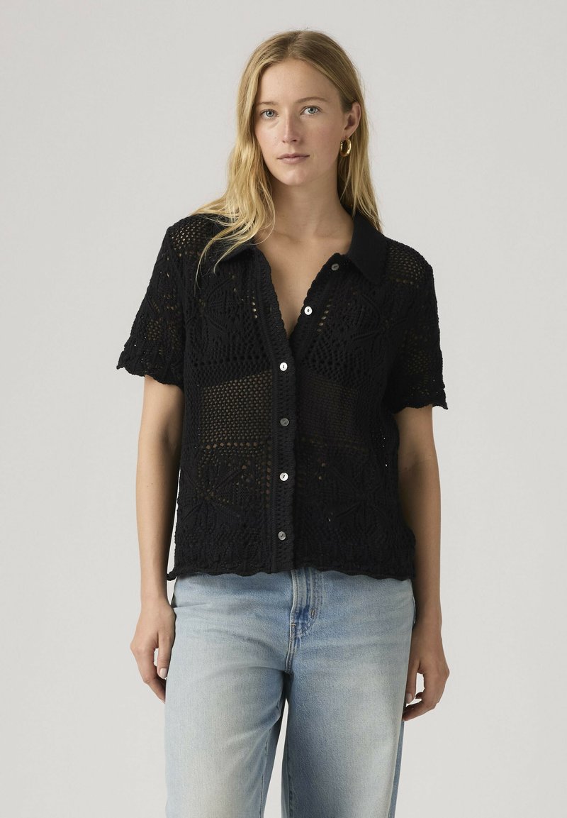 Vrouw draagt een zwart gehaakt kortermet overhemd met knopen en lichtblauwe spijkerbroek, staand tegen een effen lichtgrijze achtergrond.