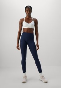 Idrottande kvinna som står vänd framåt i vit sport-bh, marinblå hög midja leggings, vita strumpor och ljusbeige sneakers på en enkel grå bakgrund.