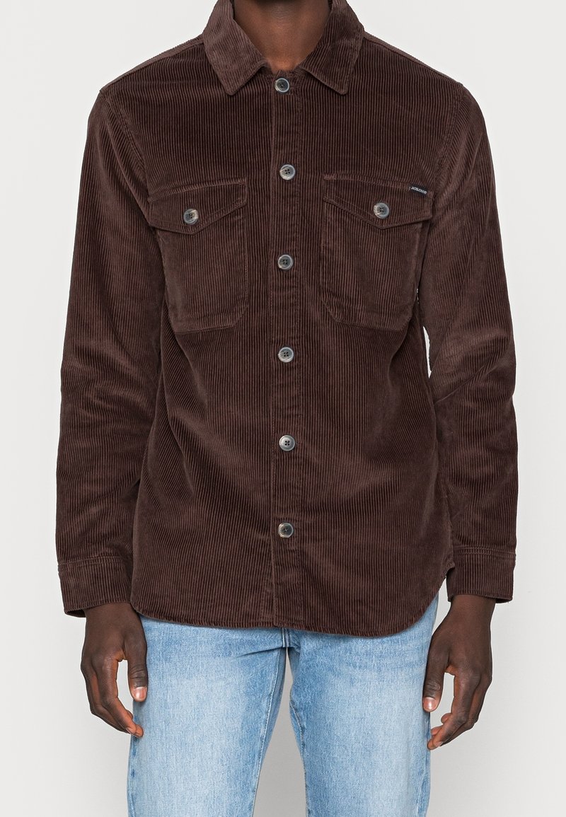 Man wearing a dark brown corduroy button-up shirt with breast pockets, paired with light blue jeans.