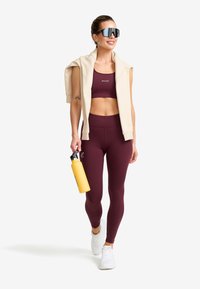 Kvinna i vinröd sport-bh och leggings med en beige sweatshirt över axlarna, hållande en gul vattenflaska, iklädd vita sneakers och svarta solglasögon.