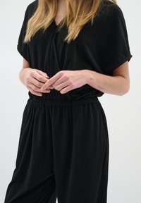 Svart jumpsuit med korta ärmar och V-ringning. Har ett elastiskt midjeband och löst sittande ben. Slät tyg, inga mönster eller accenter.