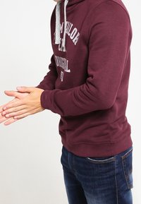 Bordeauxrode hoodie met een trekkoord, voorzien van een grijze accent en logotekst. Zachte stof, geribbelde manchetten en een ontspannen pasvorm, gecombineerd met spijkerbroek.