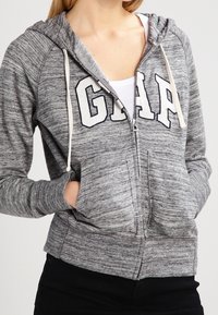 Felpa grigia con zip e cappuccio regolabile, dotata di un grande logo "GAP", tasche frontali e tessuto testurizzato.