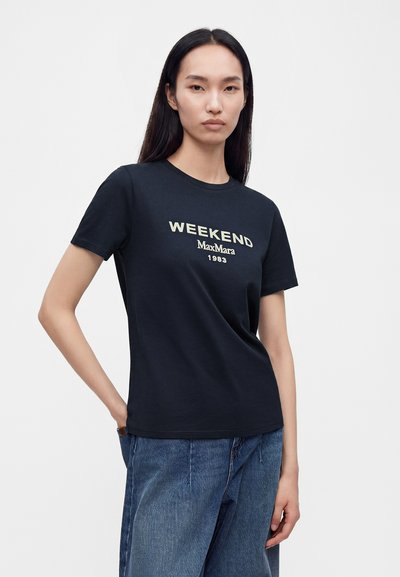 Jeune femme aux longs cheveux foncés portant un t-shirt noir avec le texte "WEEKEND MaxMara 1983" et un jean bleu sur fond blanc.