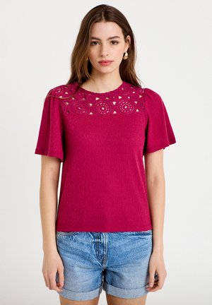 Ung kvinde iført en fuchsia top med hæklede detaljer og pufærmer, kombineret med rullede denimbukser på en hvid baggrund.