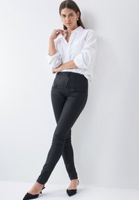 Zwarte high-waisted skinny jeans met voorzakken, gecombineerd met een wit overhemd met knopen. Model poseert tegen een effen witte achtergrond.