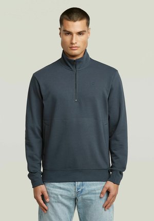 Junger Mann, der einen dunkelgrauen Pullover mit Viertel-Reißverschluss und hellblaue Jeans trägt, steht vor einem schlichten hellen Hintergrund.