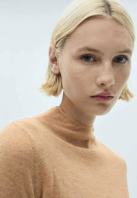 Korte mouwen, hoge hals trui van lichtoranje stof. Zachte textuur met een lichte glans. Model heeft blond haar en minimale make-up.