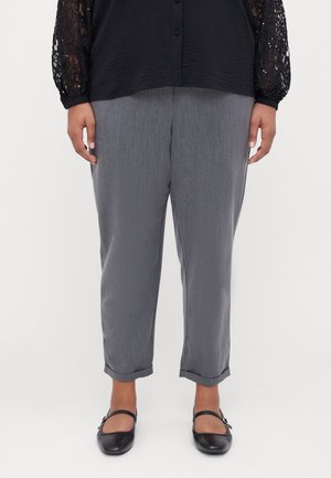 Persona che indossa pantaloni grigi risvoltati, top nero in pizzo a maniche lunghe e scarpe nere Mary Jane, in piedi su sfondo bianco.