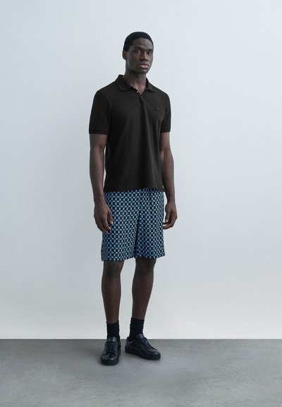 Νέος άνδρας που στέκεται, φοράει μαύρο κοντομάνικο πόλο μπλουζάκι, μπλε σορτς με σχέδια, μαύρες κάλτσες και μαύρα αθλητικά παπούτσια μπροστά σε έναν απλό τοίχο.