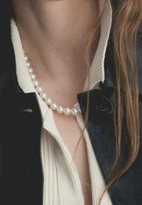 Naszyjnik z pereł okrągłych, białych noszony na lekkiej, teksturowanej bluzce i pod czarną marynarką z kołnierzem, ukazujący gładkie i lśniące powierzchnie.