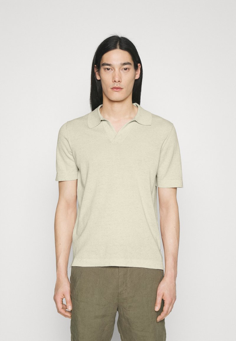 Homme aux longs cheveux noirs portant une chemise beige à manches courtes avec col et un pantalon vert olive, debout devant un fond blanc uni.