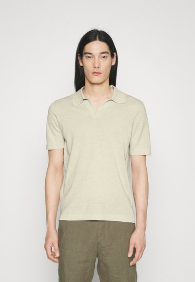 Uomo con capelli lunghi e neri che indossa una camicia beige a maniche corte con colletto e pantaloni verde oliva, in piedi contro uno sfondo bianco semplice.