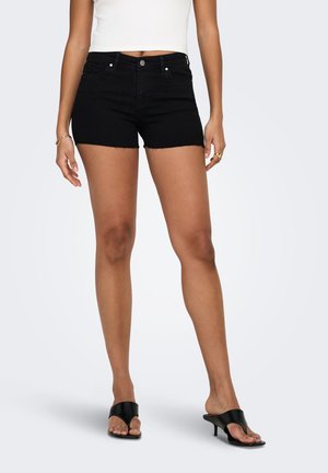 Femme portant un short en jean noir, un top blanc sans manches, des sandales noires à talons ainsi que des bagues et un bracelet en or, debout sur un fond neutre.