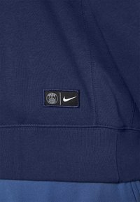 Una felpa blu navy con una texture liscia e un orlo azzurro chiaro a contrasto. Include il logo del Paris Saint-Germain e il simbolo Nike.