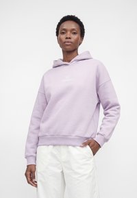 Femme portant un sweat à capuche violet clair et un pantalon blanc, debout avec une main dans la poche, sur un fond blanc uni.