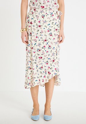 Kobieta w kremowej, midi spódnicy kopertowej w kwiaty w kolorach różowym, niebieskim i zielonym, do tego niebieskie buty na szpilce z ostrym noskiem oraz złota bransoletka.