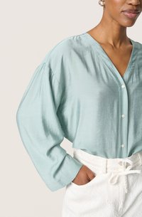 Lyseblå, silkeagtig bluse med V-hals og knapper, der har løse, lange ærmer. Parret med teksturerede, hvide bukser med snører i taljen.