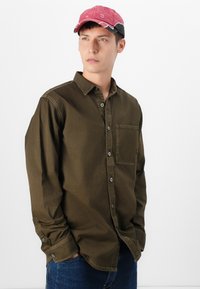Chemise olive à manches longues avec des fermetures à boutons et une poche poitrine. Accompagnée d'une casquette rose avec des accents gris et des coutures contrastées.