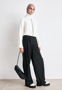 Cremfarbener, gerippter Rollkragenpullover mit schwarzen Nähdetails, kombiniert mit schwarzen, weit geschnittenen Hosen und klobigen, zweifarbigen Plateauschuhen. Schwarze Tasche.