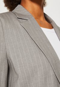 Blazer gris de raya diplomática, tela ligera, solapa afilada, hombros estructurados, bolsillo de un solo pecho, superpuesto a una blusa blanca.