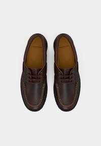 Um par de sapatos de barco de couro castanho-escuro, com atacadores, detalhes costurados e solas pretas, vistos de cima sobre um fundo branco.