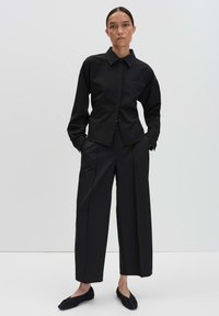 Vrouw staat met handen in de zakken, gekleed in een zwarte overhemd met knopen, brede zwarte broek en zwarte platte schoenen tegen een effen achtergrond.