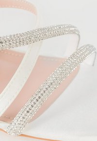 Sandale en satin blanc avec des brides entrecroisées ornées de strass. La semelle intérieure lisse rose et la semelle plate créent un design élégant.