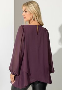 Blusa chiffon prugna con maniche lunghe e gonfie, schiena drappeggiata e dettaglio a occhio di chiave al collo, abbinata a pantaloni neri aderenti.