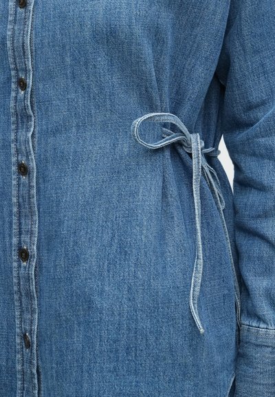 Πουκάμισο από τζιν σε ανοιχτό μπλε χρώμα, με κουμπιά μπροστά και ζώνη στη μέση, με απαλή υφή και ορατή ραφή.