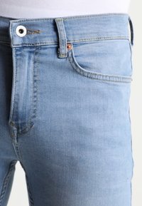 Vaaleansiniset farkut, joissa on näkyvä etutasku, vyölenkki ja metallinen nappisuljenta vyötärö- ja reiden yläosassa.