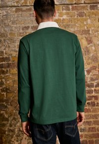 Chemise verte à manches longues avec un col blanc, en coton ; présente une texture lisse et une coupe droite, portée sur un jean noir.