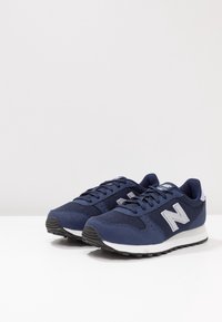 Marinblå sportskor tillverkade av mesh och syntetmaterial, med en vit gummisula och en reflekterande logotyp på sidan.