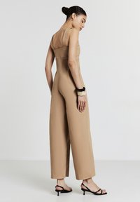Frau in einem ärmellosen beigen Jumpsuit, schwarzen Riemchen-High-Heels und klobigen Armbändern, die seitlich vor einem schlichten weißen Hintergrund steht.