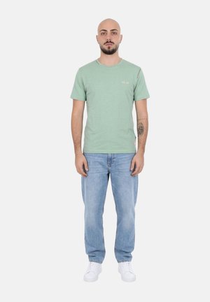 Uomo con barba e tatuaggio in piedi rivolto frontalmente, indossa una t-shirt verde, jeans azzurri e scarpe da ginnastica bianche su uno sfondo uniforme.