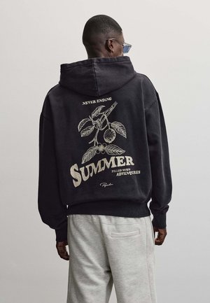 Mann trägt schwarzen Hoodie mit botanischem Design und dem Text „Never Ending Summer Filled With Adventures“ auf dem Rücken, kombiniert mit hellgrauen Hosen.