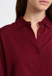 Bordeaux knopenoverhemd met een col, vijf knoopsluitingen en een gestructureerde stof, met een klein logo geborduurd nabij de kraag.