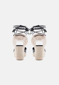 Ζευγάρι πλατφόρμες με τακούνια από πλεγμένο σχοινί, υφαντό μπεζ ύφασμα και μαύρα κορδόνια στον αστράγαλο, εμφανιζόμενα από πίσω σε λευκό φόντο.