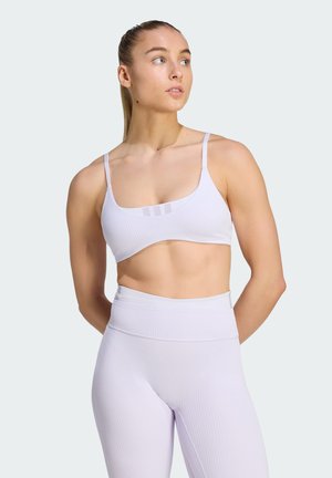 Frau, die mit den Händen hinter dem Rücken steht, einen weißen gerippten Sport-BH und hochtaillierte Leggings trägt und nach links blickt vor einfachem Hintergrund.